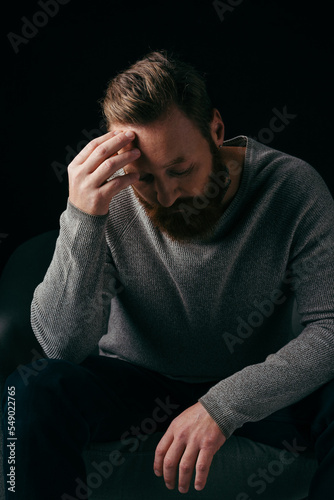 Bearded man in jumper touching forehead while standing near armchair isolated on black.