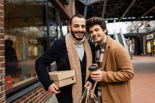happy and fashionable gay couple with purchases and coffee to go looking at camera near showcase on street.