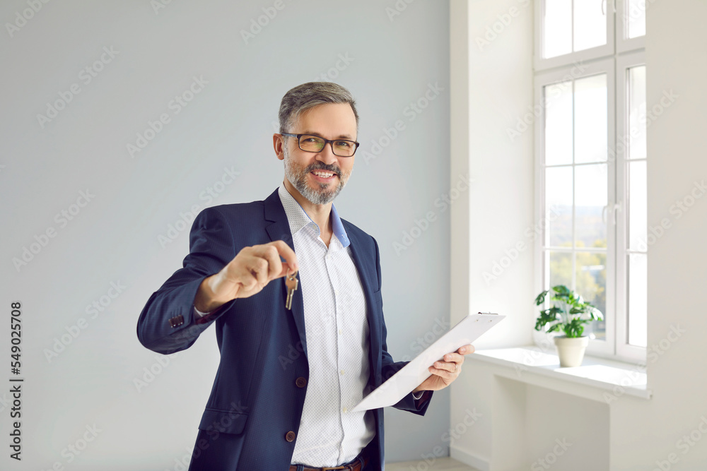 Portrait of friendly male real estate agent showing key to house that you can buy or rent. Smiling Caucasian middle-aged man in business suit posing with clipboard and key in bright room. Portrait of friendly male real estate agent showing key to house that you can buy or rent. Smiling Caucasian middle-aged man in business suit posing with clipboard and key in bright room.
