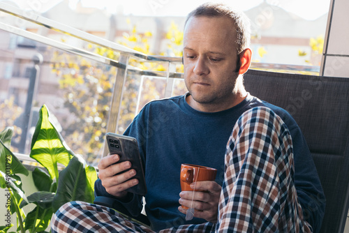 young man looking at his cell phone and drinking tea or infusion on the terrace of his home