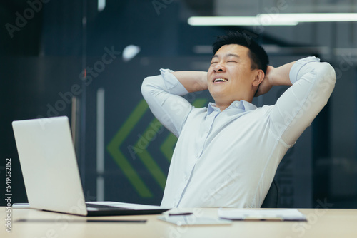 A young handsome Asian man, a businessman sits in the office at the desk. He holds his hands behind his head, closes his eyes, and smiles. Satisfied with his work, rests during working hours.