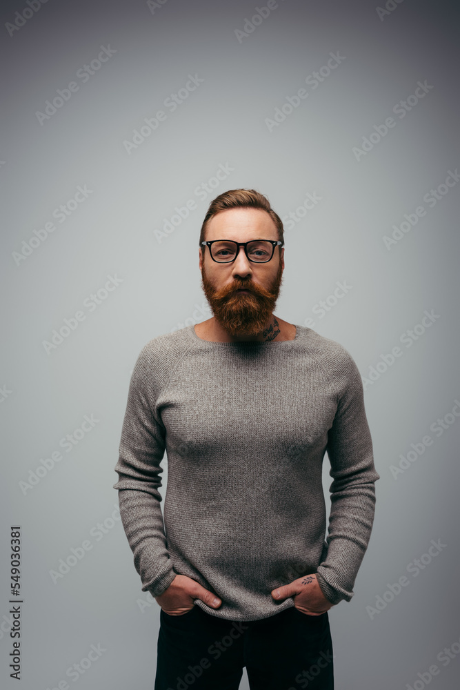 Stylish bearded man in jumper holding hands in pockets of pants isolated on grey. Stylish bearded man in jumper holding hands in pockets of pants isolated on grey.