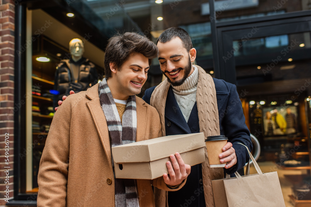 smiling bearded man with coffee to go looking at shoebox in hand of boyfriend near blurred showcase on street. smiling bearded man with coffee to go looking at shoebox in hand of boyfriend near blurred showcase on street.