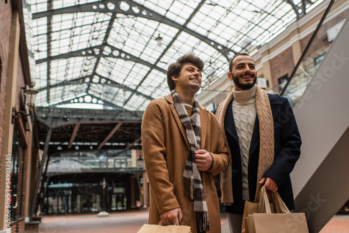 cheerful gay boyfriends in trendy clothes standing with shopping bags under transparent roof on street.