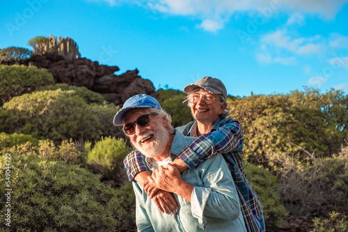 Happy caucasian senior couple enjoying travel and nature in mountain range. Man carries piggyback his wife