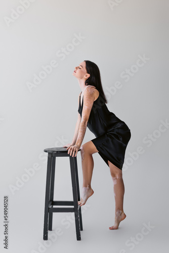 Side view of young model with vitiligo posing near chair on grey background.