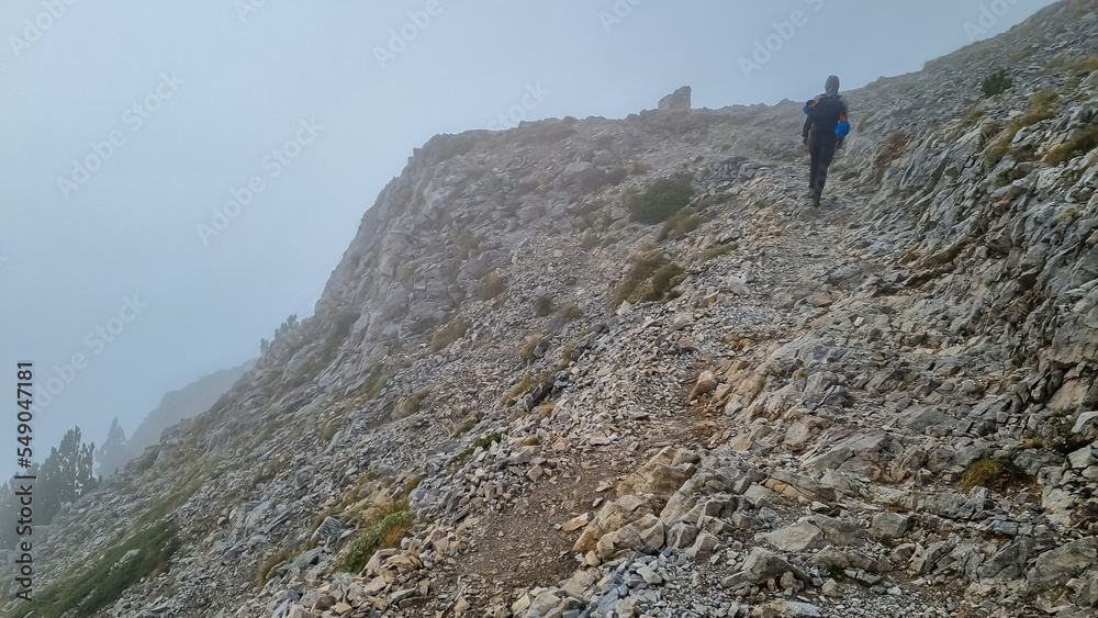 Man trekking on mystical foggy hiking trail leading to Mount Olympus (Mytikas, Skala, Stefani) in Mt Olympus National Park, Thessaly, Greece, Europe. Scenic view of cloud covered slopes and ridges