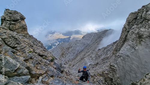 Man climbing on mystical foggy hiking trail leading to Mount Olympus (Mytikas, Skala, Stefani) in Mt Olympus National Park, Macedonia, Greece, Europe. Scenic view of cloud covered slopes and ridges