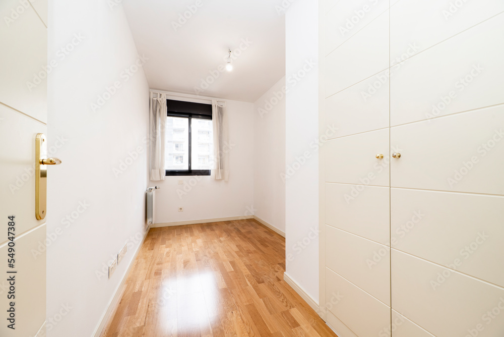Empty small bedroom with white wooden built-in wardrobe doors, black anodized aluminum windows and white painted walls and wooden floors Empty small bedroom with white wooden built-in wardrobe doors, black anodized aluminum windows and white painted walls and wooden floors