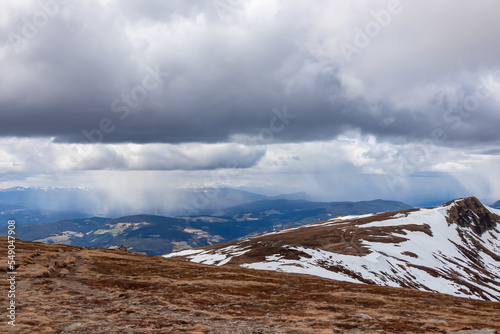 Panoramic view on summit cross of mountain peak Gertrusk seen from Ladinger Spitz  Saualpe  Carinthia  Austria  Europe. Snow covered hiking trail over alpine meadows and hills. Cloudy early spring day