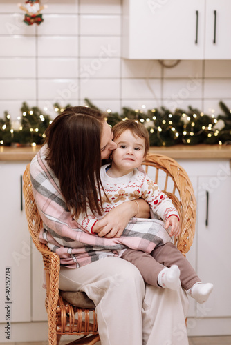 Merry Christmas and Happy New Years Holidays. Young mother hugging and kissing her baby daughter, sitting and relaxing together in wicker chair at kitchen decorated for Xmas. Time of family party.