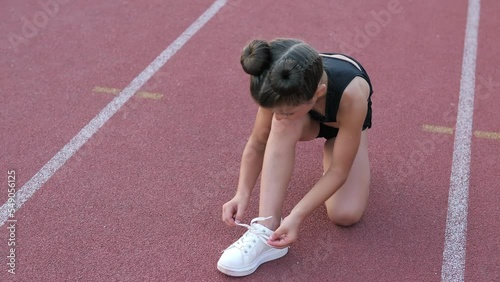 A little girl is tying her shoelaces on the running ground, preparing for competitions. A sporty lifestyle. Preparing for an athletic training on a beautiful day at the stadium. Sport, athletics.