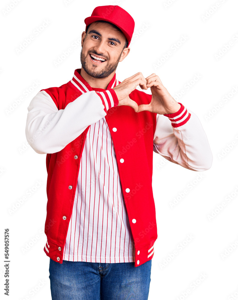 Young handsome man wearing baseball jacket and cap smiling in love showing heart symbol and shape with hands. romantic concept. Young handsome man wearing baseball jacket and cap smiling in love showing heart symbol and shape with hands. romantic concept.