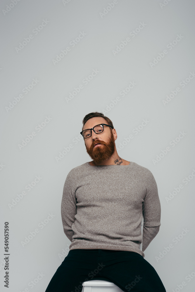 Trendy bearded man in eyeglasses posing on chair isolated on grey. Trendy bearded man in eyeglasses posing on chair isolated on grey.