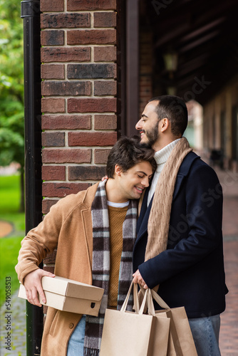 happy gay man with braces and closed eyes holding shoebox while leaning on bearded boyfriend with shopping bags.