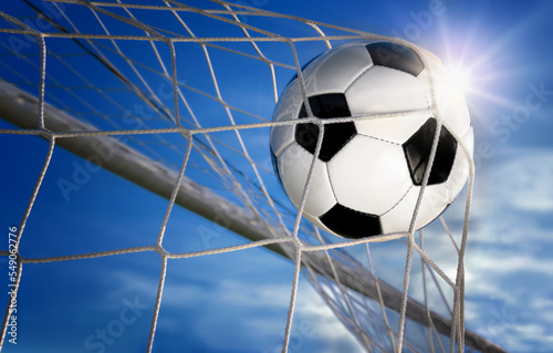 Football or soccer goal, a ball with classic design flying into the net, with blue sky and sun in the background