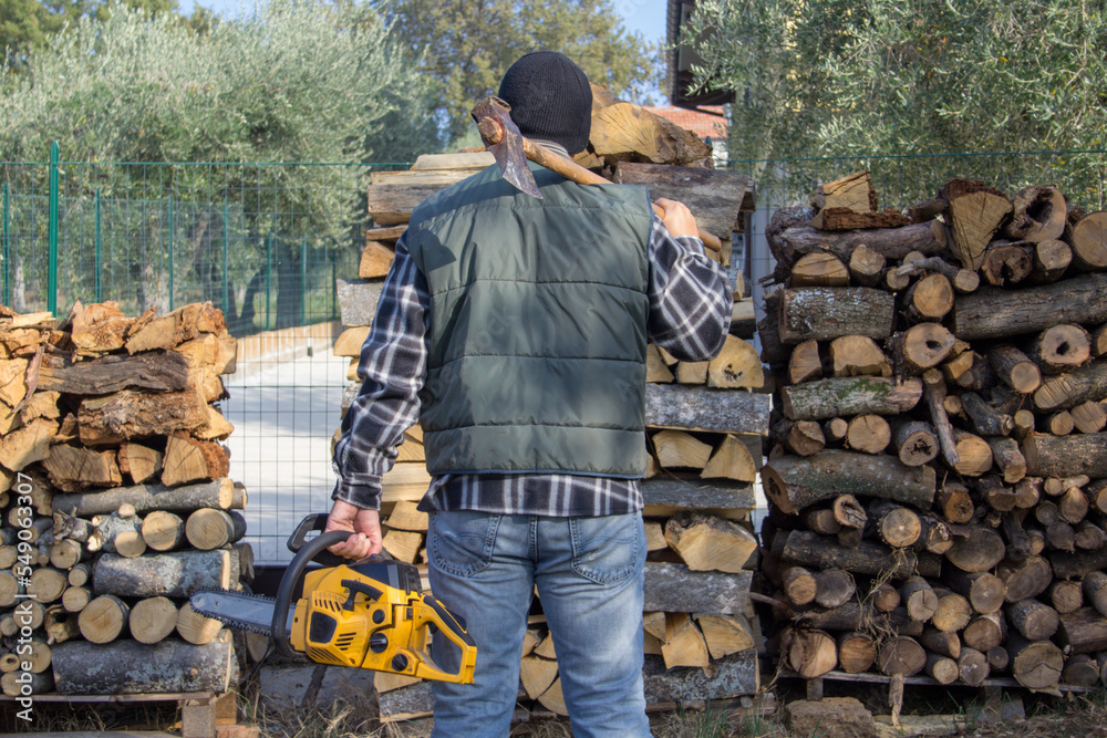 Image of a lumberjack from behind with ax and chainsaw in hand after compiling a pile of wood as preparation for winter. Heating with the home fireplace
