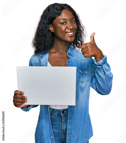 Beautiful african young woman holding blank empty banner smiling happy and positive, thumb up doing excellent and approval sign