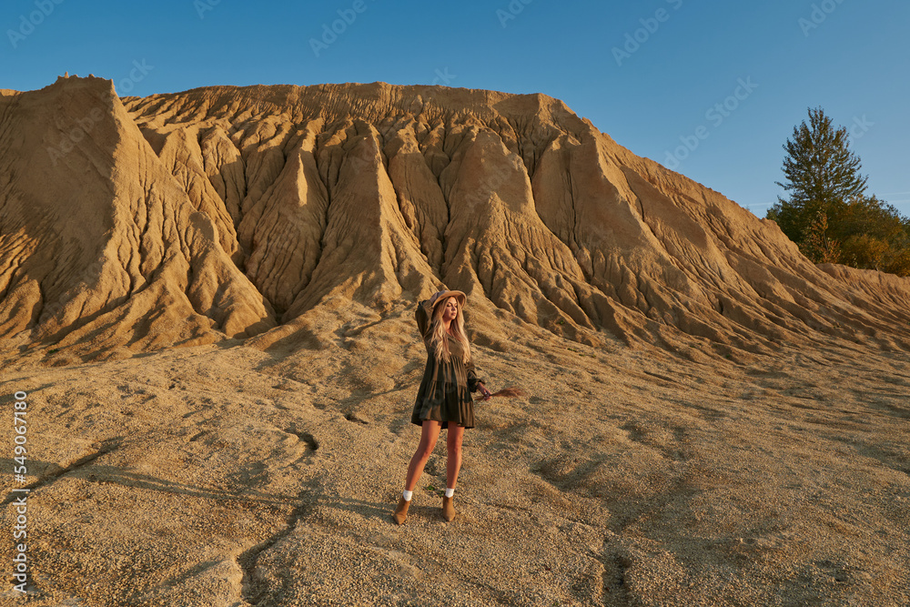 a beautiful young woman in a dress and a hat with dry grass in her hands on the background of a huge sandy mountain and a beautiful clear blue sky. travel, beautiful nature