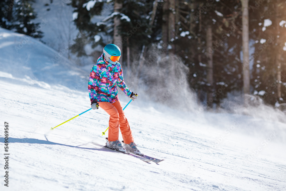 Young skier in bright clothes sliding down the slope on a sunny day at a mountain resort.