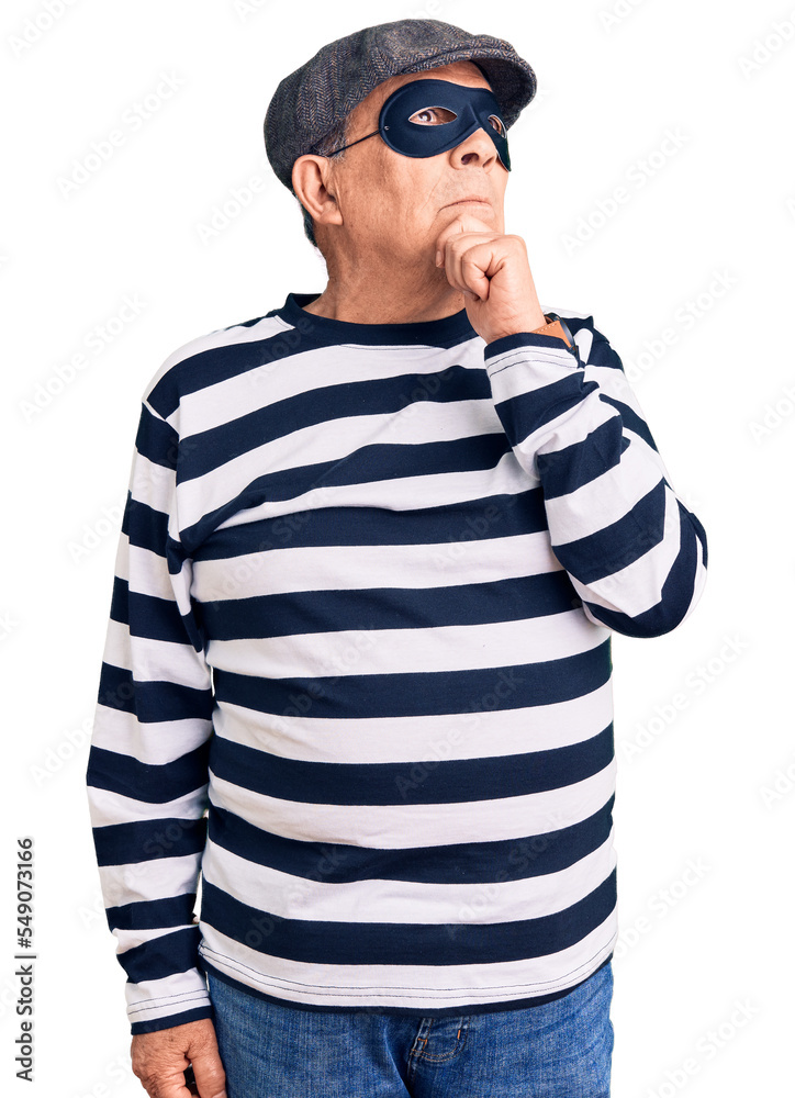Senior handsome man wearing burglar mask and t-shirt with hand on chin thinking about question, pensive expression. smiling with thoughtful face. doubt concept. Senior handsome man wearing burglar mask and t-shirt with hand on chin thinking about question, pensive expression. smiling with thoughtful face. doubt concept.