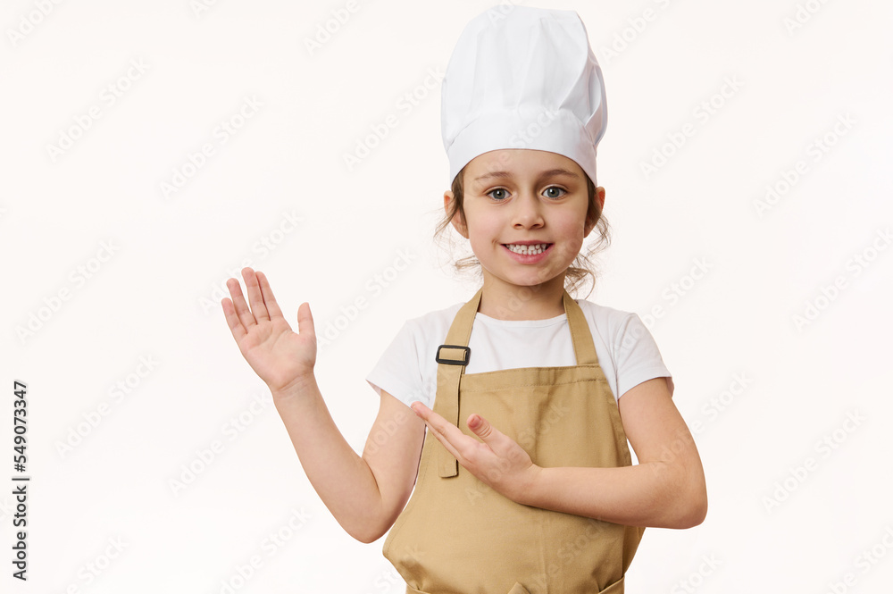 Charming little girl in chef's uniform, smiling sweetly at camera, pointing her hand at a blank place for advertising text, isolated on a white background. Concept for cooking class. Food consumerism Charming little girl in chef's uniform, smiling sweetly at camera, pointing her hand at a blank place for advertising text, isolated on a white background. Concept for cooking class. Food consumerism