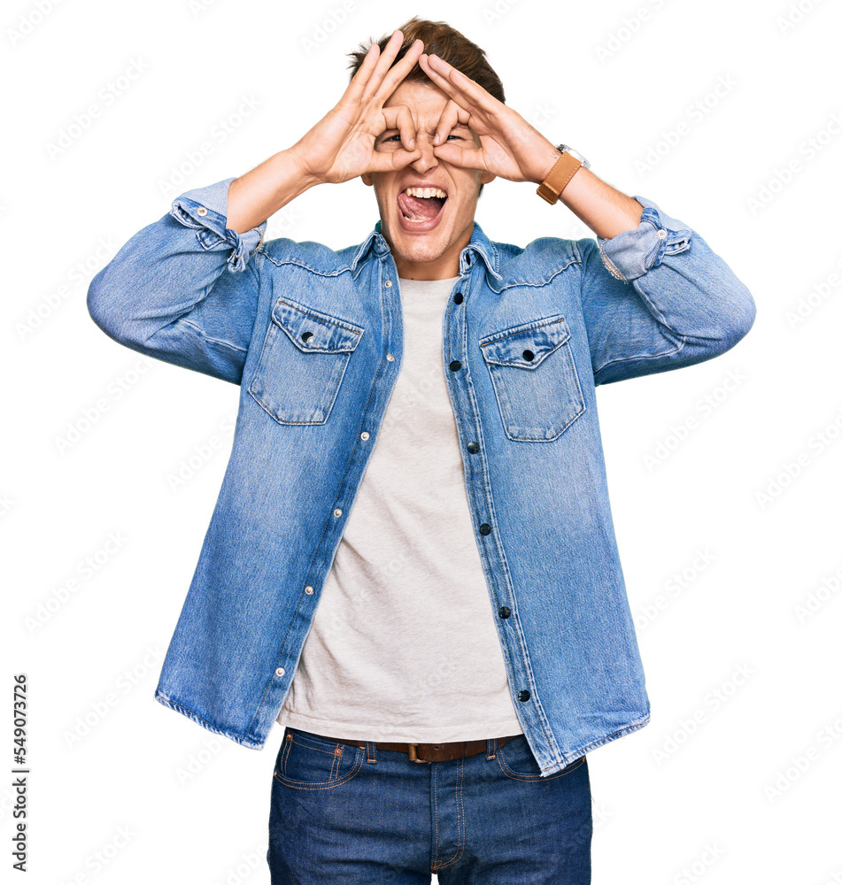 Handsome caucasian man wearing casual denim jacket doing ok gesture like binoculars sticking tongue out, eyes looking through fingers. crazy expression.