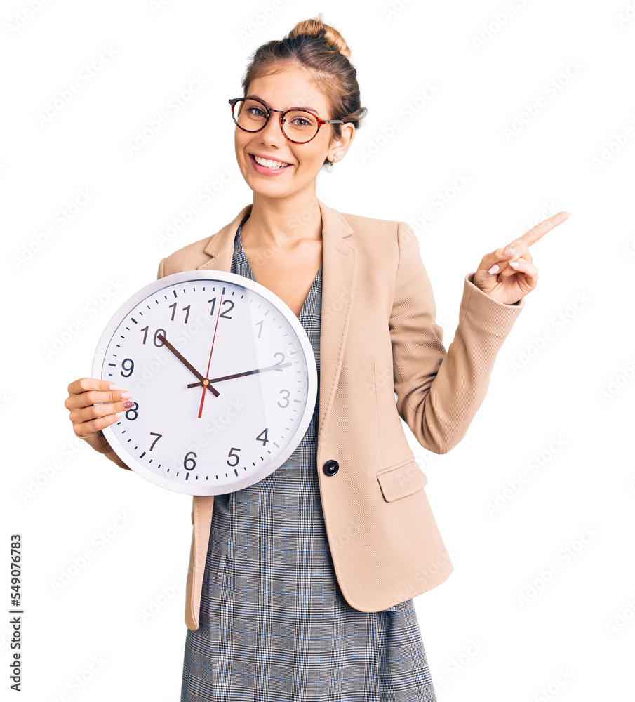 Beautiful caucasian woman with blonde hair wearing business jacket and holding clock smiling happy pointing with hand and finger to the side Beautiful caucasian woman with blonde hair wearing business jacket and holding clock smiling happy pointing with hand and finger to the side