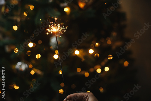 Fototapeta Hand holding firework against christmas tree lights in dark room