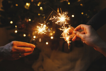 Hands holding fireworks against christmas lights in dark room. Happy New Year! Atmospheric holiday. Friends celebrating with burning sparklers in hands on background of stylish illuminated tree