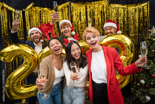 Interracial businesswomen holding champagne near businessmen and balloons in shape of 2023 numbers in office