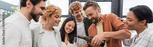 Photography cheerful businessman pouring champagne near smiling interracial colleagues havin