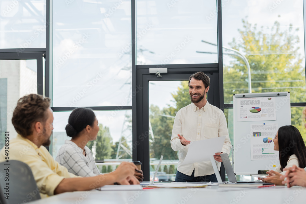 Fototapeta premium smiling businessman with document pointing with hand while talking to multiethnic team in meeting room