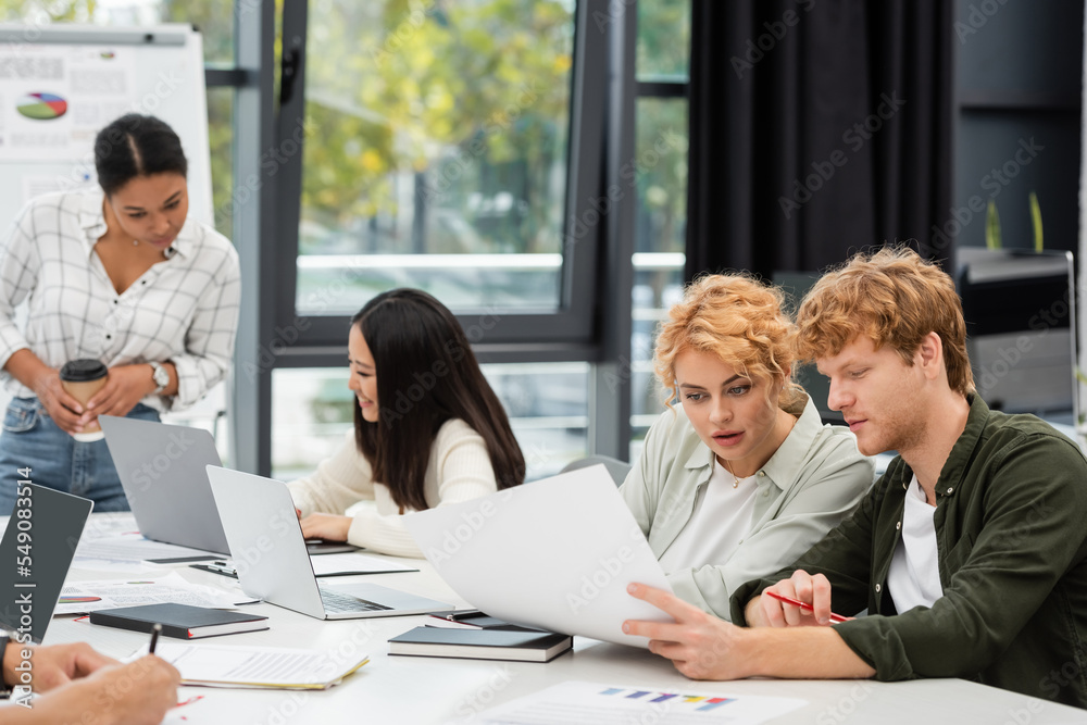 young managers looking at document near multiethnic coworkers on blurred background young managers looking at document near multiethnic coworkers on blurred background