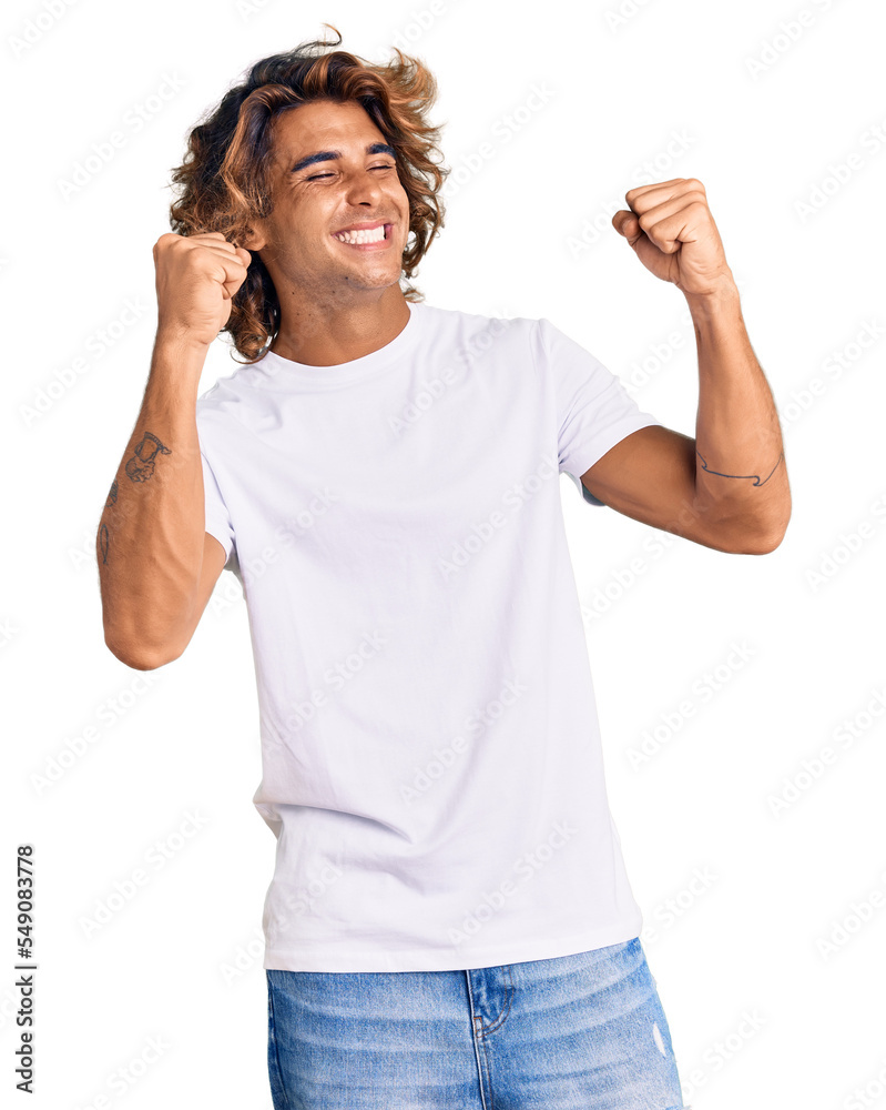 Young hispanic man wearing casual white tshirt very happy and excited doing winner gesture with arms raised, smiling and screaming for success. celebration concept. Young hispanic man wearing casual white tshirt very happy and excited doing winner gesture with arms raised, smiling and screaming for success. celebration concept.