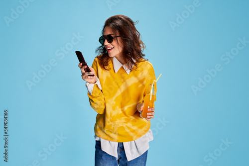 Portrait of young beautiful girl texting on phone and drinking juice isolated over blue background