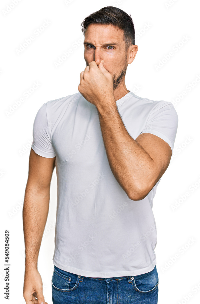 Handsome man with beard wearing casual white t shirt smelling something stinky and disgusting, intolerable smell, holding breath with fingers on nose. bad smell Handsome man with beard wearing casual white t shirt smelling something stinky and disgusting, intolerable smell, holding breath with fingers on nose. bad smell