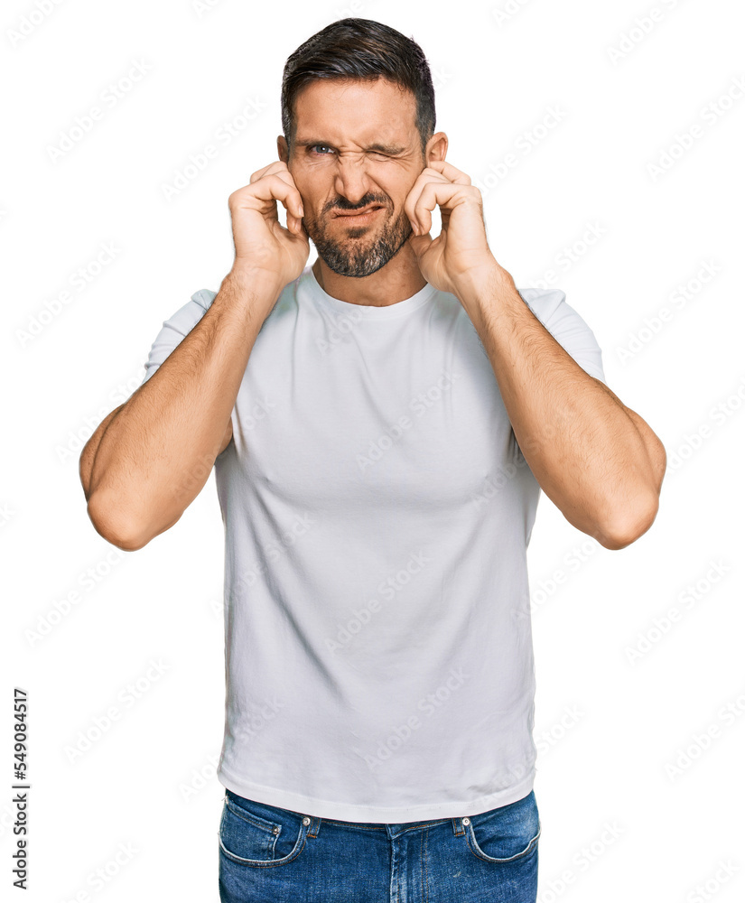 Handsome man with beard wearing casual white t shirt covering ears with fingers with annoyed expression for the noise of loud music. deaf concept. Handsome man with beard wearing casual white t shirt covering ears with fingers with annoyed expression for the noise of loud music. deaf concept.