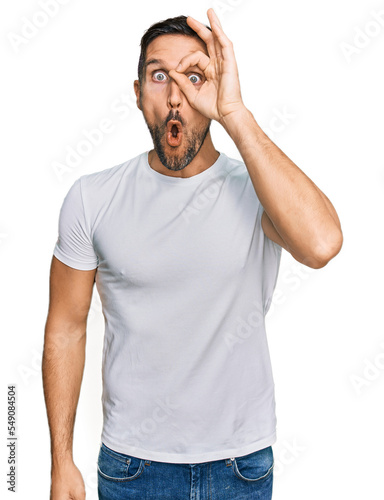 Handsome man with beard wearing casual white t shirt doing ok gesture shocked with surprised face, eye looking through fingers. unbelieving expression.