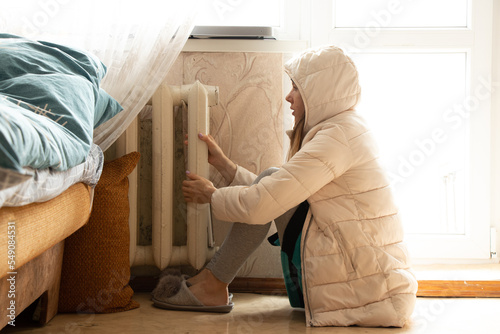 A girl in a winter jacket is sitting near a cold battery in an apartment in Ukraine, there is no heat due to shelling in Ukraine, a cold house