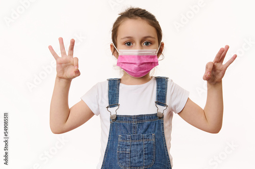 Adorable Caucasian little girl wearing pink protective medical mask, gestures with hands, shows OK signs, looks at camera, isolated over white background. Free space. Pediatrics. Health care. Medicine photo