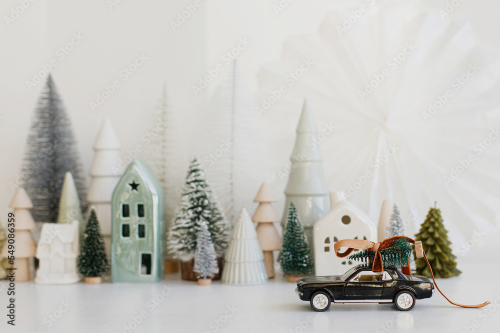 Merry Christmas and Happy Holidays! Stylish little car carrying christmas tree on background of Christmas miniature snowy village. Festive winter scene on white table, holiday banner