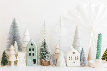 Merry Christmas and Happy Holidays! Modern christmas scene, miniature cozy snowy village. Stylish little Christmas trees and houses decorations on white table. Winter banner, scandinavian decor