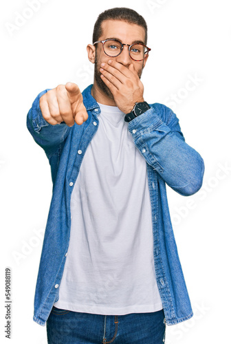 Young caucasian man wearing casual clothes laughing at you, pointing finger to the camera with hand over mouth, shame expression