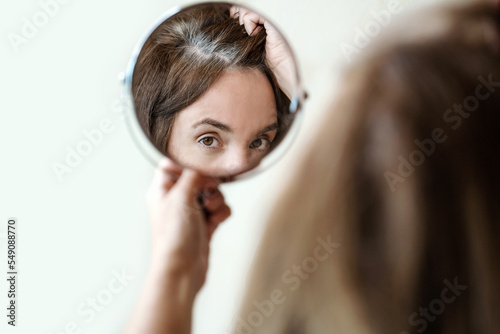 Close-up of a mirror with a reflection of a woman's face. Young woman is examining her gray hair roots