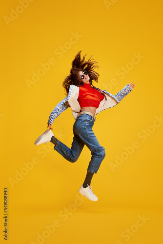 Portrait of young beautiful girl posing in stylish casual clothes, cheerfully jumping isolated over yellow background