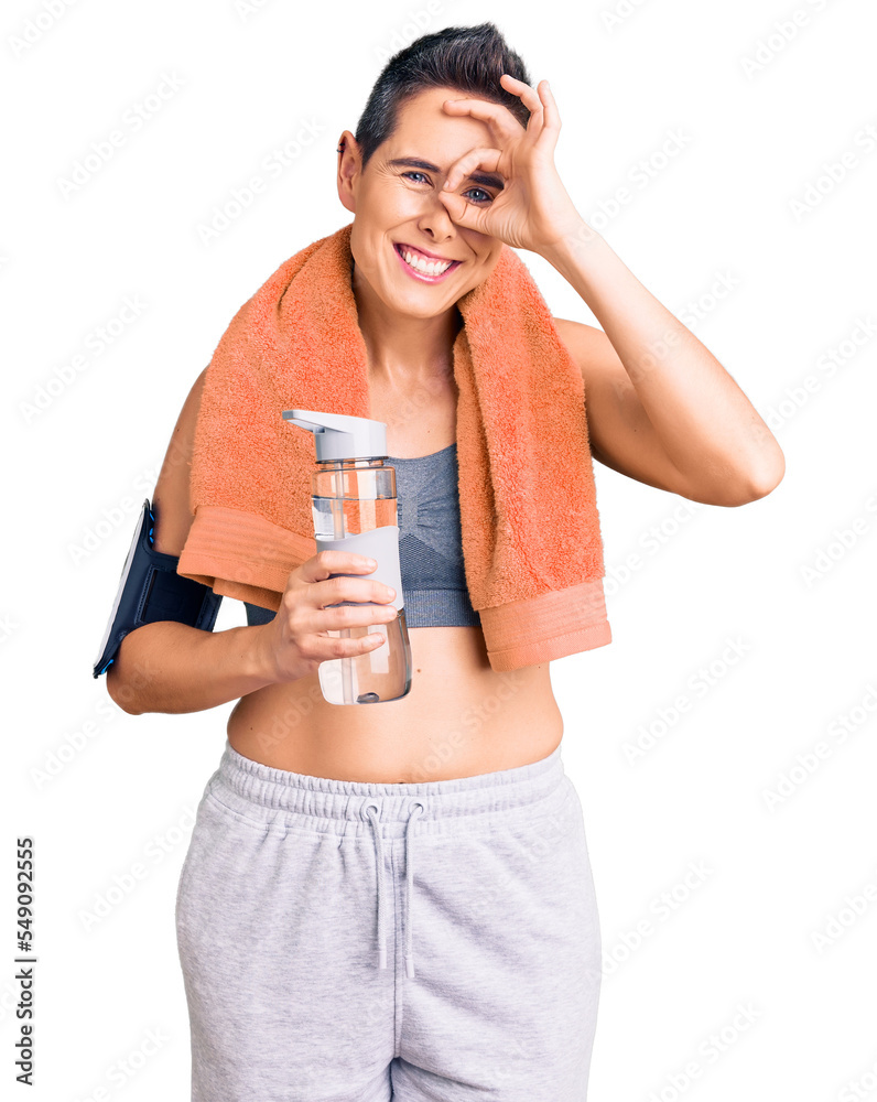 Young woman with short hair wearing sportswear and towel holding bottle of water smiling happy doing ok sign with hand on eye looking through fingers Young woman with short hair wearing sportswear and towel holding bottle of water smiling happy doing ok sign with hand on eye looking through fingers