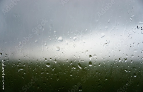Glasscheibe mit Regentropfenmuster vor grünem Feld und weißgrauem Himmel bei Regen am Nachmittag im Winter