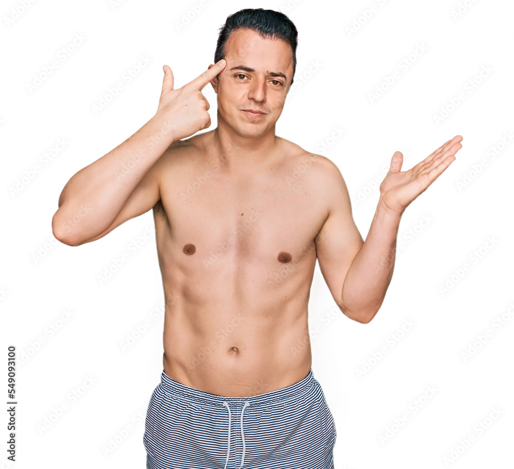 Handsome young man wearing swimwear shirtless confused and annoyed with open palm showing copy space and pointing finger to forehead. think about it. Handsome young man wearing swimwear shirtless confused and annoyed with open palm showing copy space and pointing finger to forehead. think about it.
