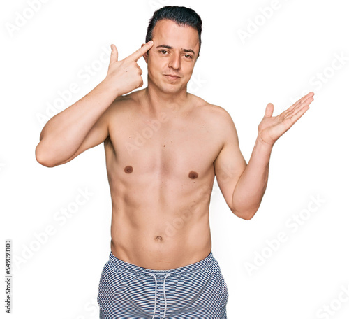 Handsome young man wearing swimwear shirtless confused and annoyed with open palm showing copy space and pointing finger to forehead. think about it.